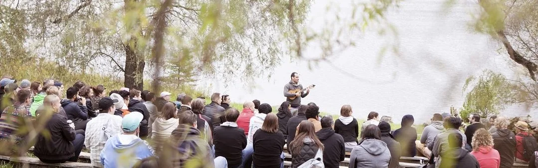 Jonathan Mann performing for an outdoor audience beside a lake